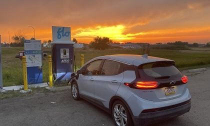 A Chevrolet Bolt EV charges at a Flo station under a vibrant sunset, with lush greenery in the background.