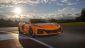 An orange Corvette stands on a racetrack, illuminated by sunlight, with dramatic clouds in the background and lush greenery in the distance.