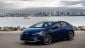 A blue Toyota Prius parked by a waterfront with city skyline in the background under a cloudy sky.