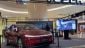 A red Tesla Model Y is showcased in a modern mall setting, with a digital screen displaying a robotic character beside it.